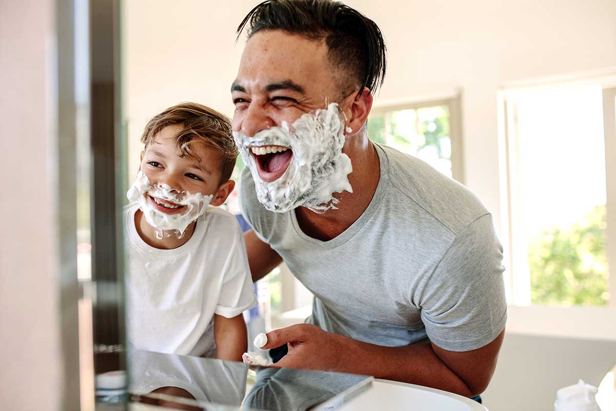 Father and son with shaving foam beards at the bathroom mirror.