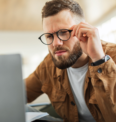 Young bearded man peering through glasses at computer with a puzzled look.