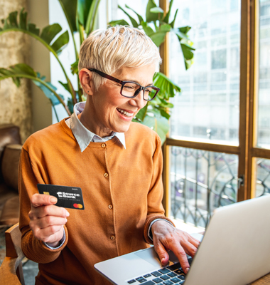 Mature woman placing an order on her laptop, holding 1st Source Bank Resource Plus Debit Mastercard.