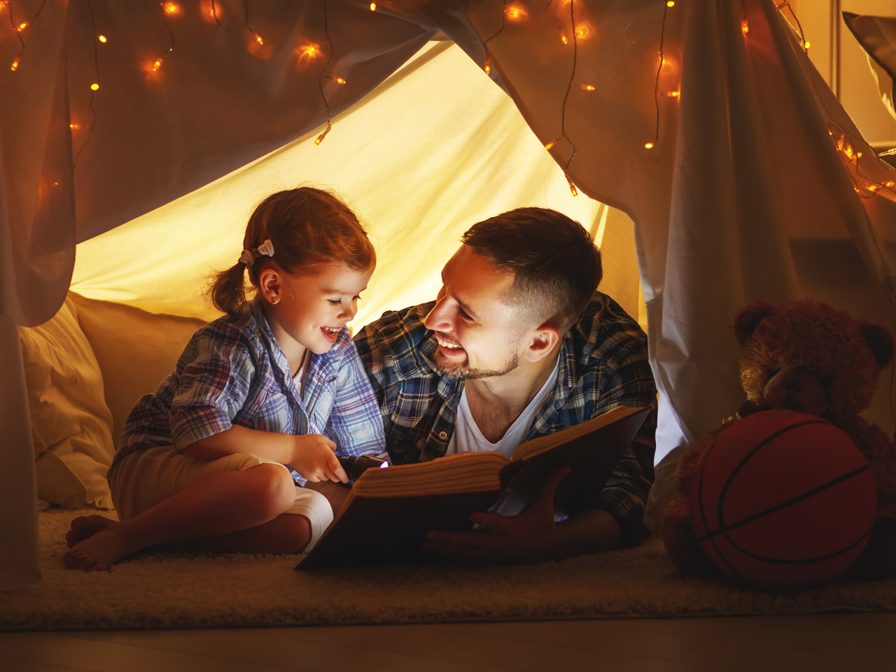 Father reading to daughter in tent with flashlight.
