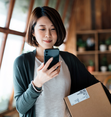 Young woman holding shipping box and looking at smartphone.