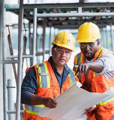 Two male construction workers looking at plans under scaffolding.