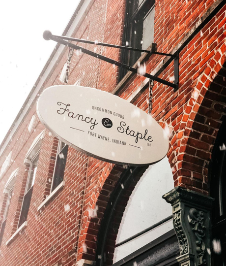 Oval Fancy & Staple sign hanging in front of brick building.