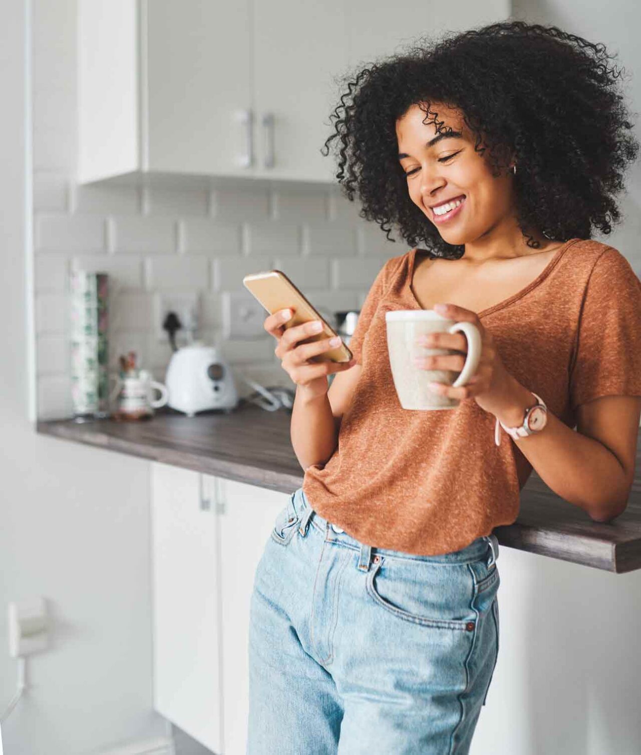 A woman with curly hair checks her online banking on her mobile phone and enjoys a cup of coffee in her kitchen