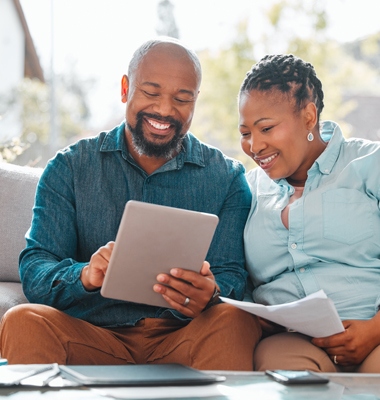 Couple sitting on living room couch paying bills on a smart tablet.