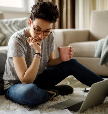 Young woman sitting on living room floor with laptop, looking over bills.