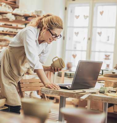 Female pottery maker using laptop in shop.