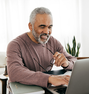 Mature gentleman holding glasses and performing calculations on a laptop.