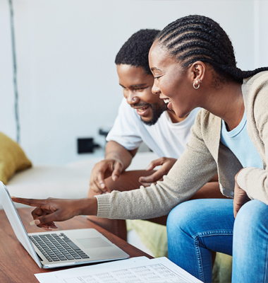 Young couple on living room couch happily reviewing savings account info on laptop.
