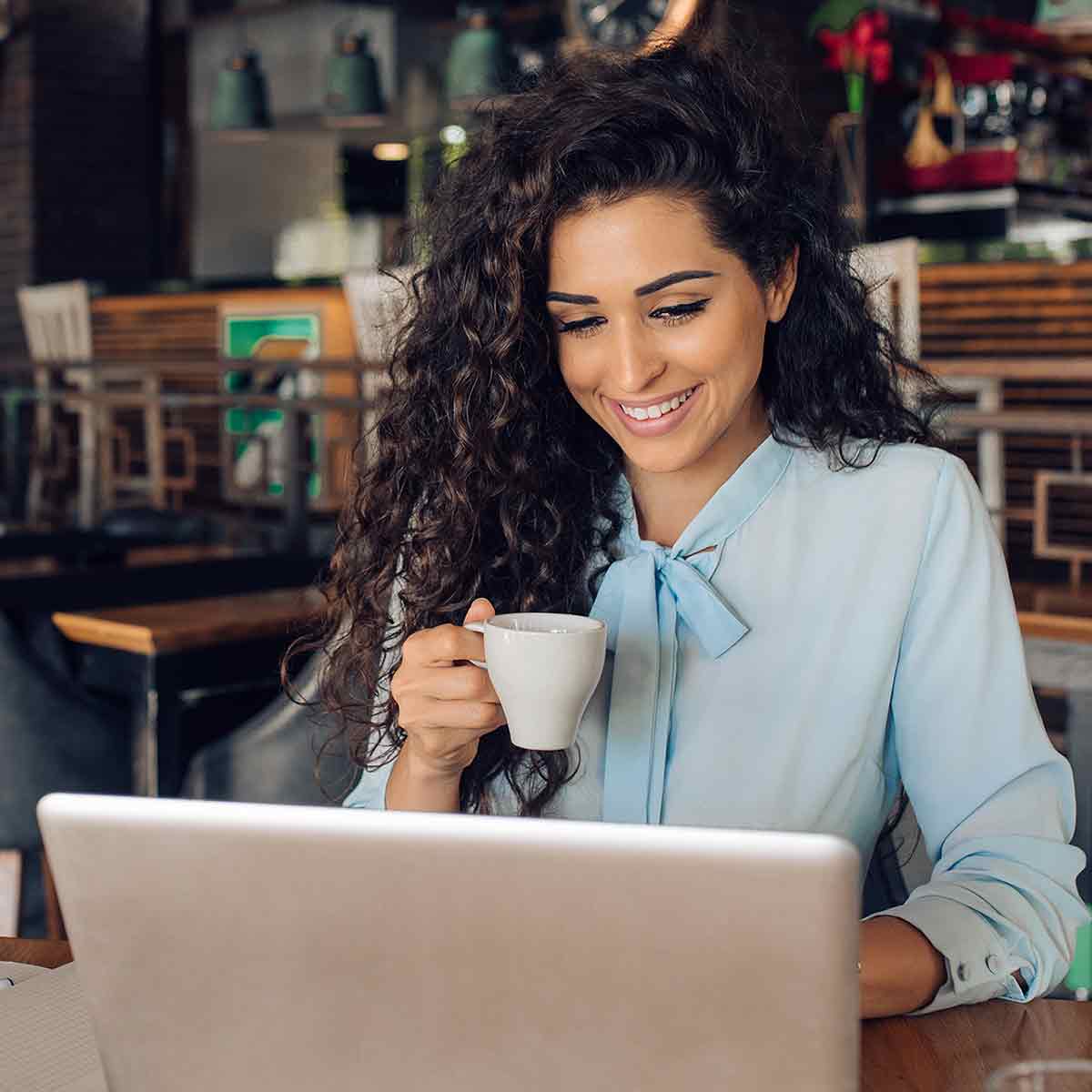 A woman with curly hair happily uses her laptop, displaying a bright smile on her face.