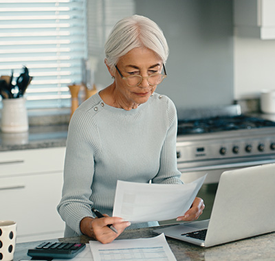 A woman sitting at a kitchen table, focused on her laptop and financial paperwork