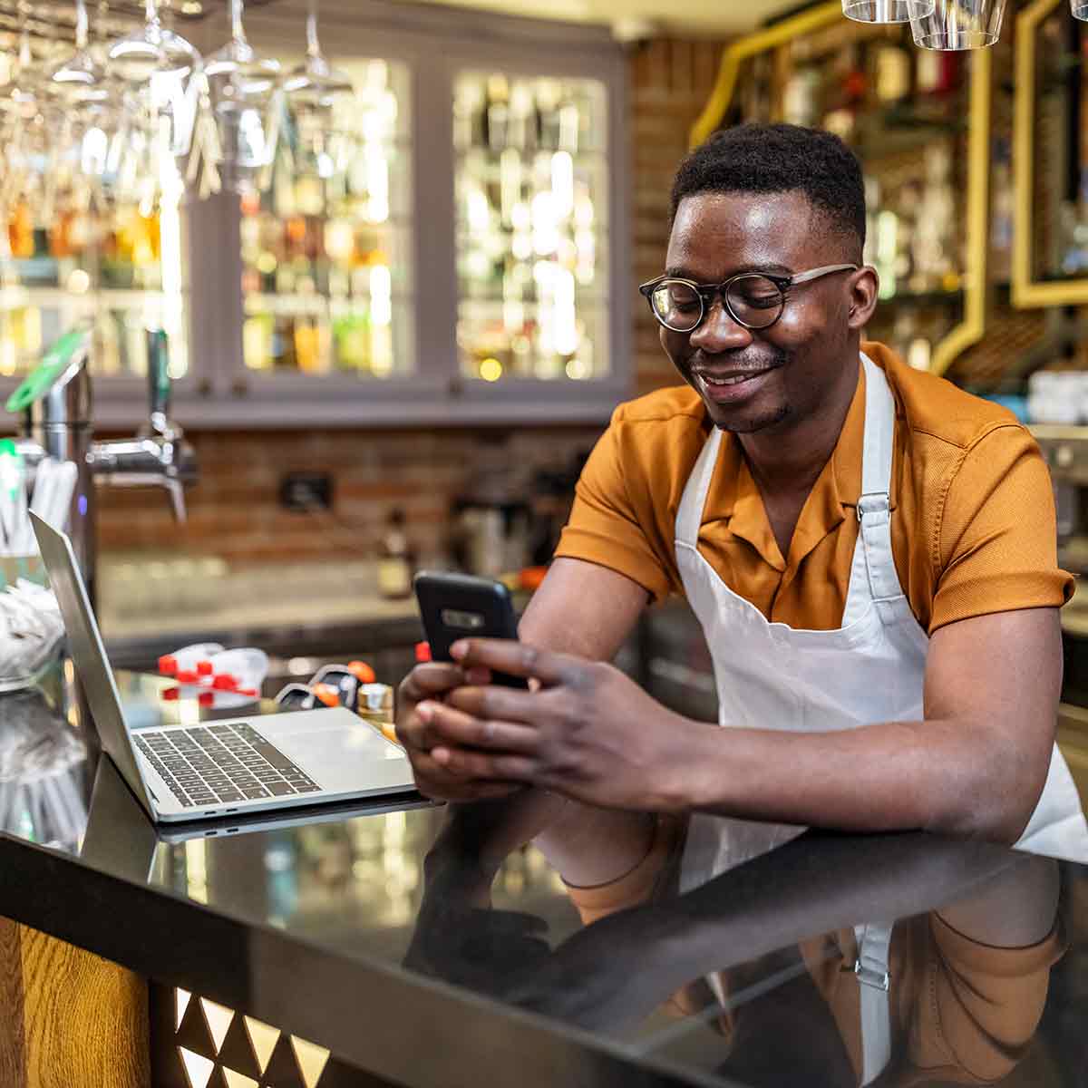 A small business owner wearing an apron grins as he checks his phone in his restaurant.