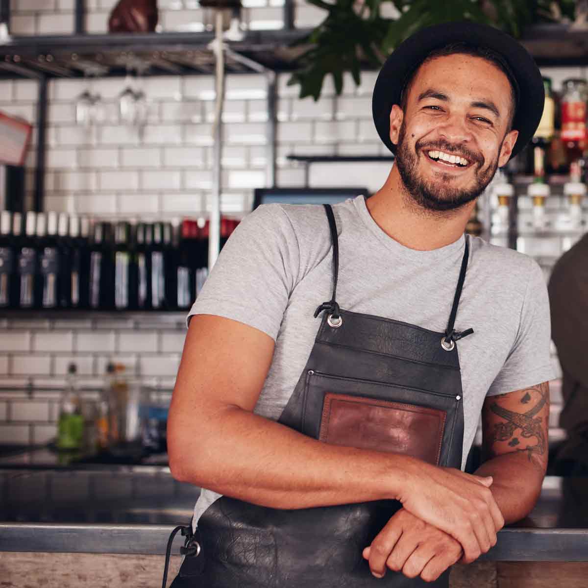 A man in an apron stands in front of a bar, smiling warmly.