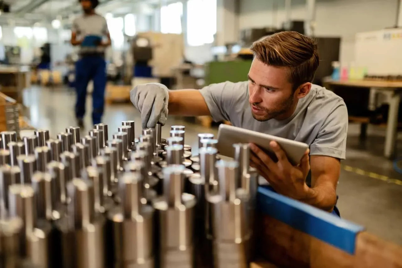 Man inspecting metal parts on an assembly line, utilizing a digital tablet for quality control in a modern manufacturing facility.