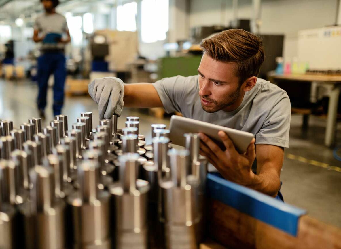 Man inspecting metal parts on an assembly line, utilizing a digital tablet for quality control in a modern manufacturing facility.