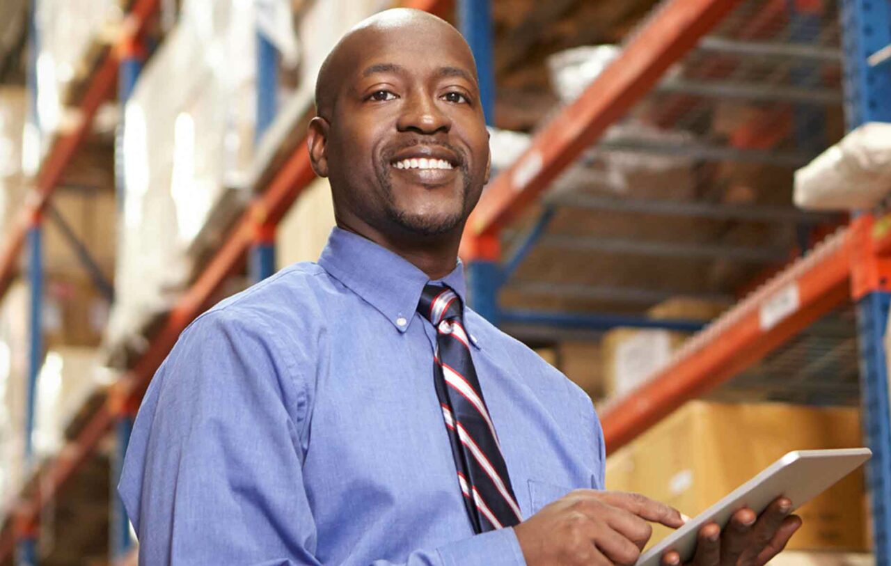 Smiling man wearing a tie, working a tablet in a warehouse.