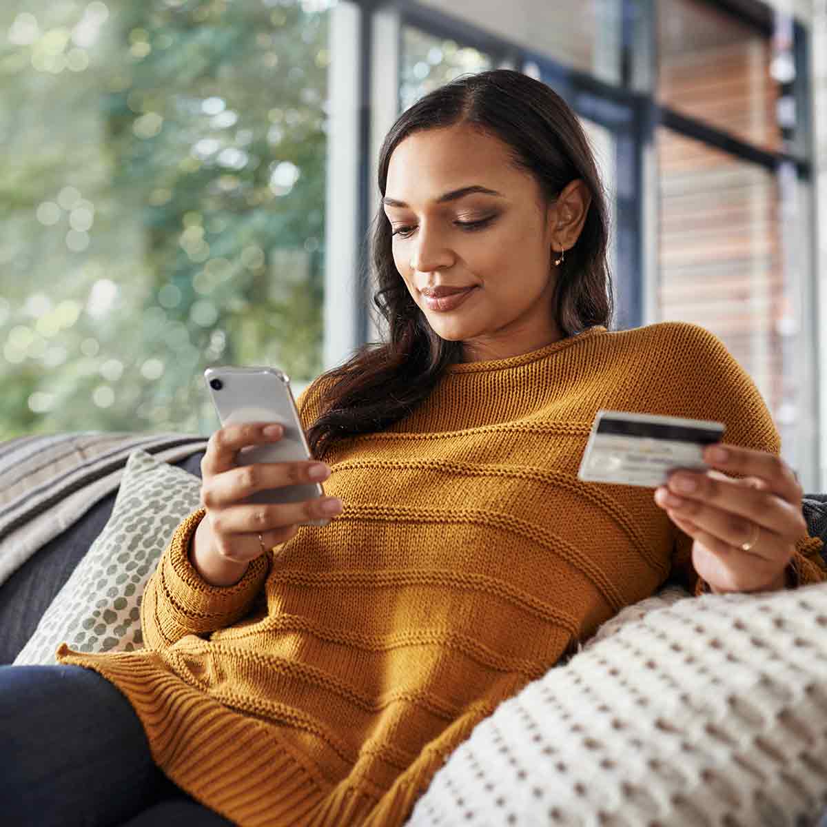 A woman reclines on her couch, using her smartphone and debit card to make a payment