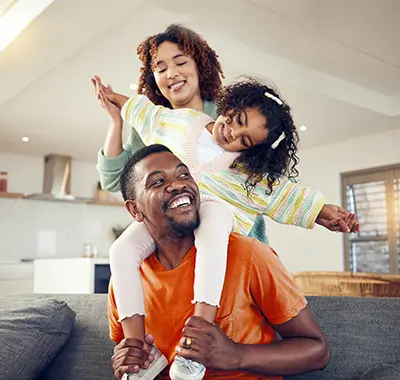Happy African American family enjoying quality time together at home. Smiling father with young daughter on his shoulders and mother standing behind, showcasing a loving and joyful family moment indoors.