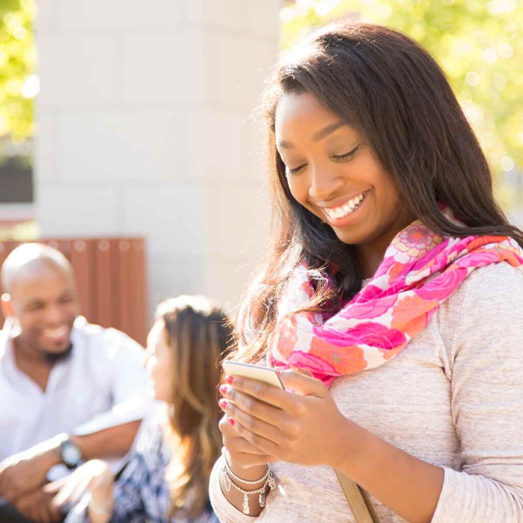 A female college student is smiling while walking through campus, looking at her cell phone.