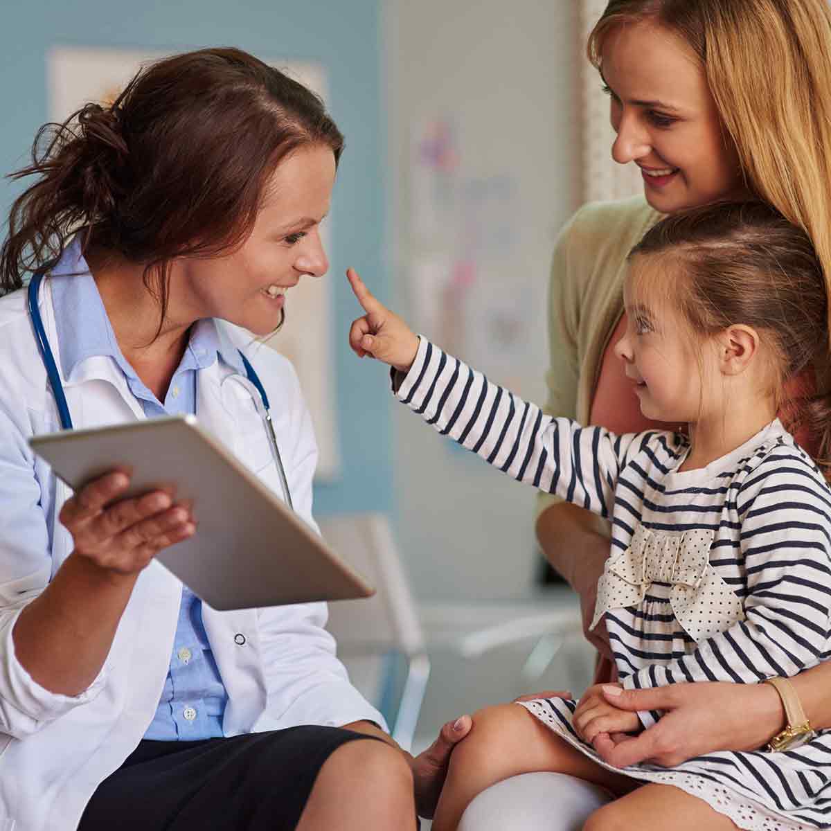 A friendly doctor smiles and plays with a curious child, who is sitting on her mother's lap.