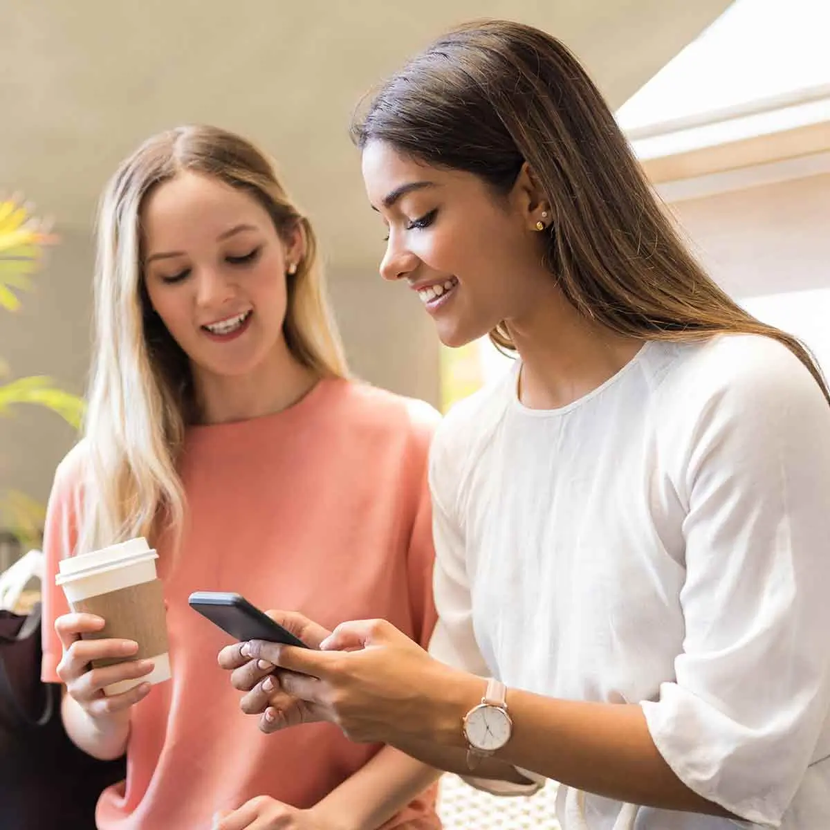 Two friends having coffee and laughing at what's displayed on one girl's phone