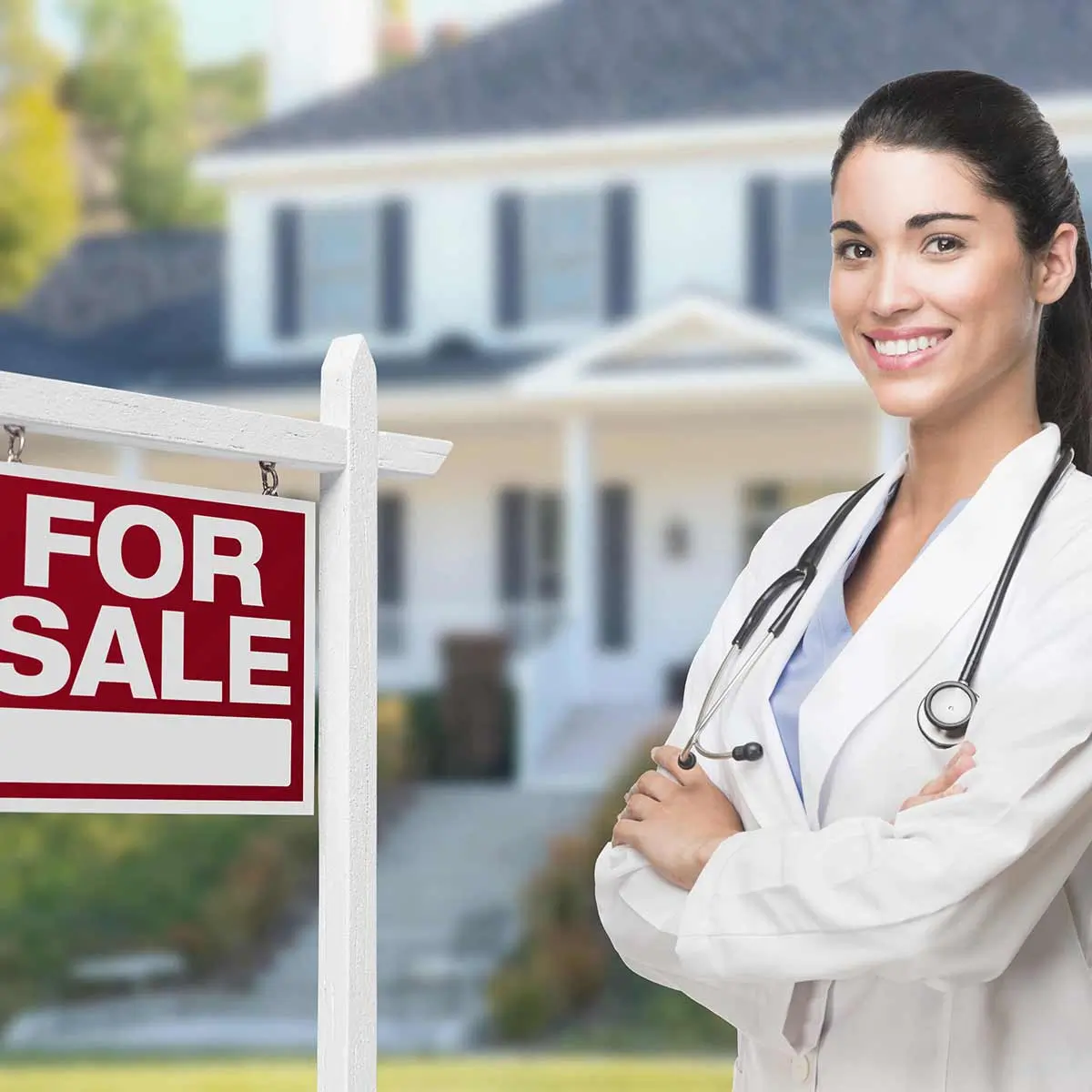 Smiling female doctor standing in front of a house with a for sale sign, representing physician home loan opportunities for medical professionals.