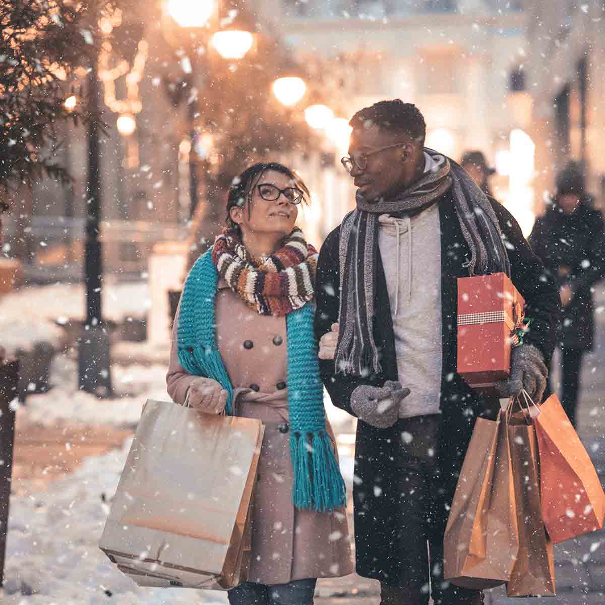 Couple Christmas shopping downtown in the evening with light snow catching the outdoor lights of streetlamps and shops.