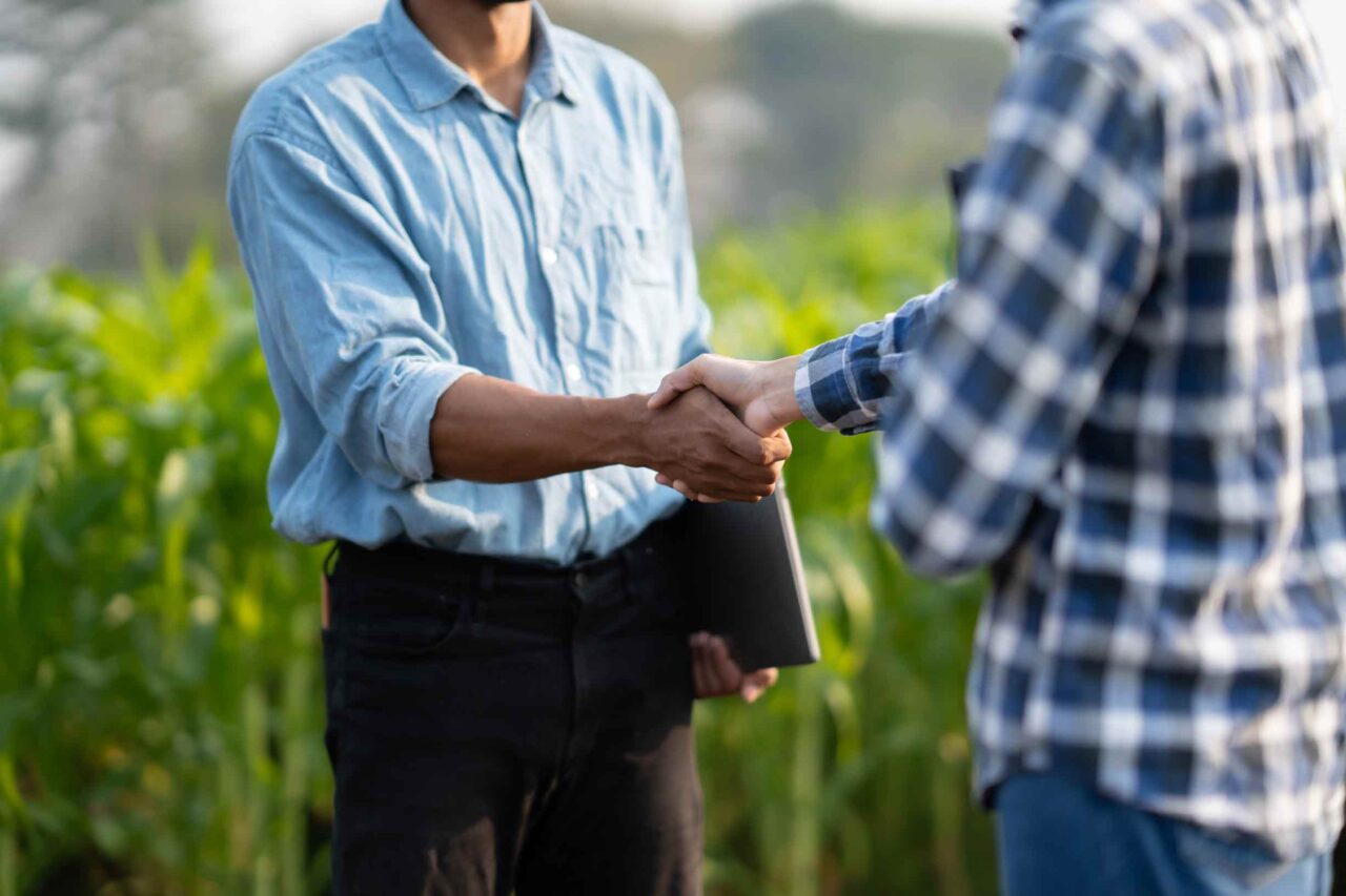 A banker and a farmer shake hands amidst a corn field, engaging in a conversation about farm loans and business banking.
