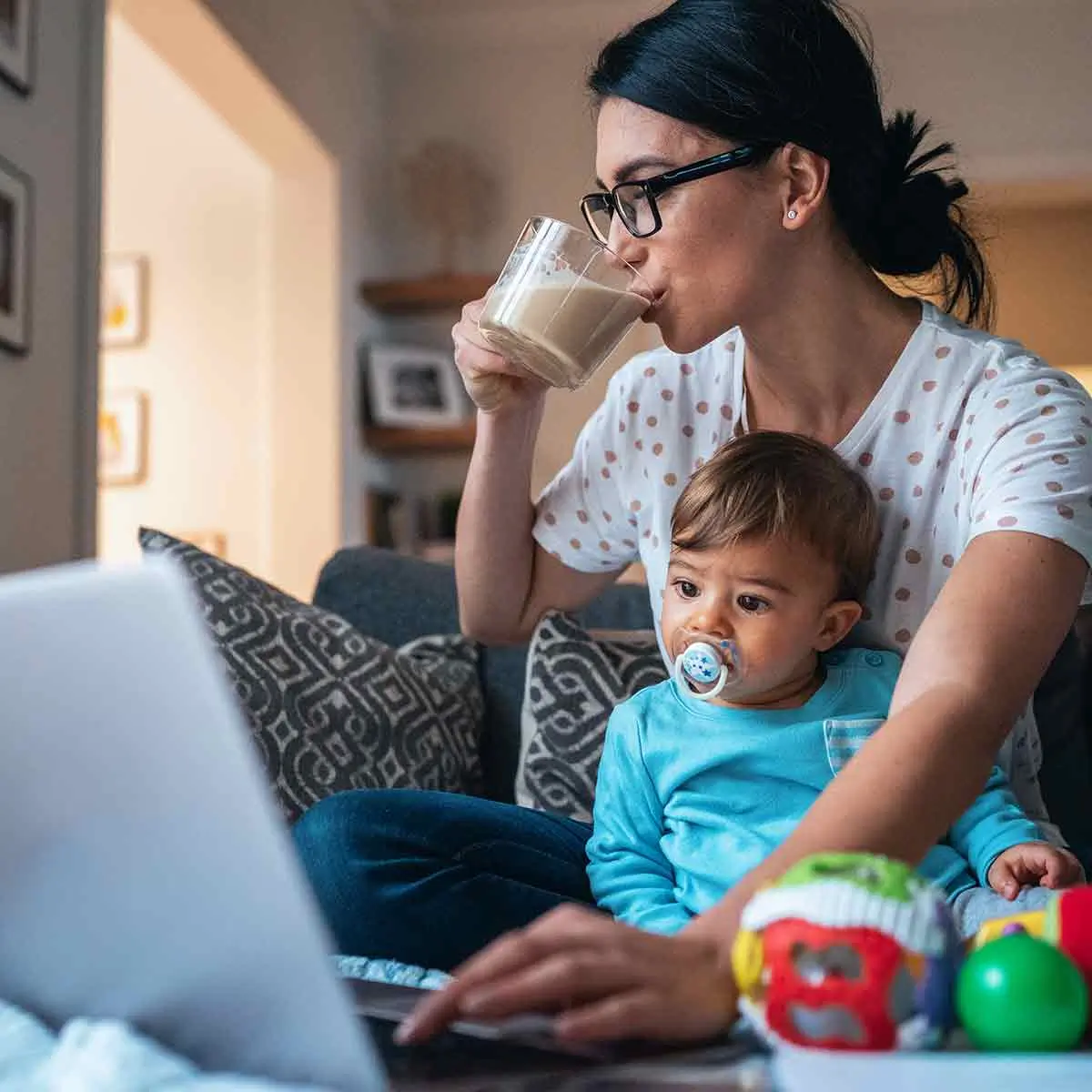 Woman working on laptop with toddler on couch, showing the challenges that working moms face