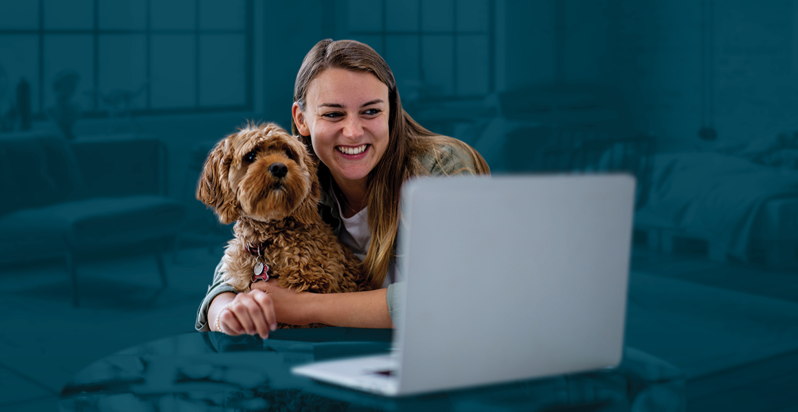 A smiling young woman hugs her dog while using her laptop
