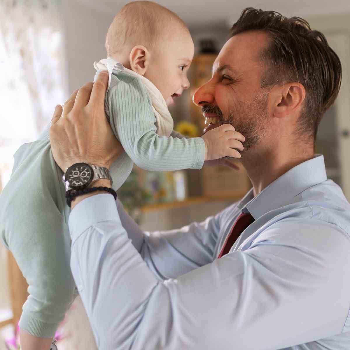 Father in business attire lifting and playing with his baby after work, symbolizing work/life balance