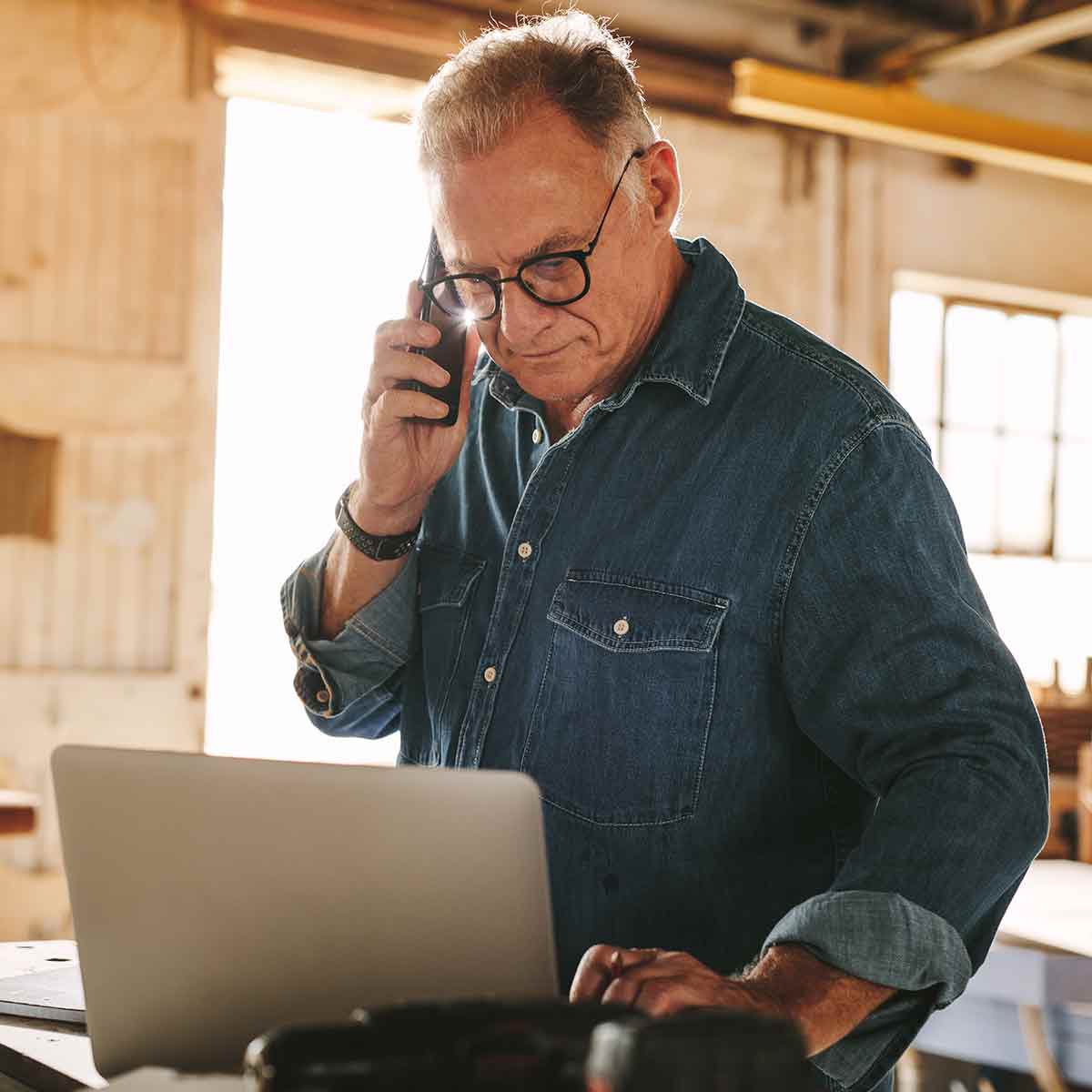Mature carpenter working on laptop and talking on cell phone