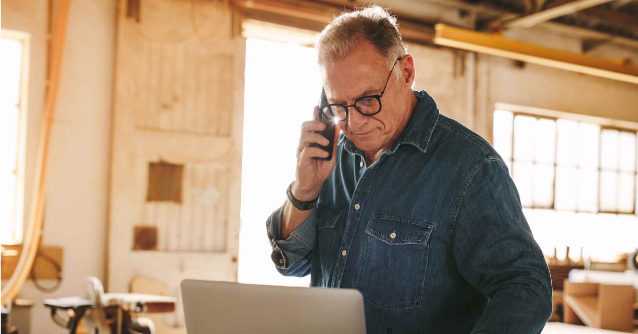Mature carpenter working on laptop and talking on cell phone
