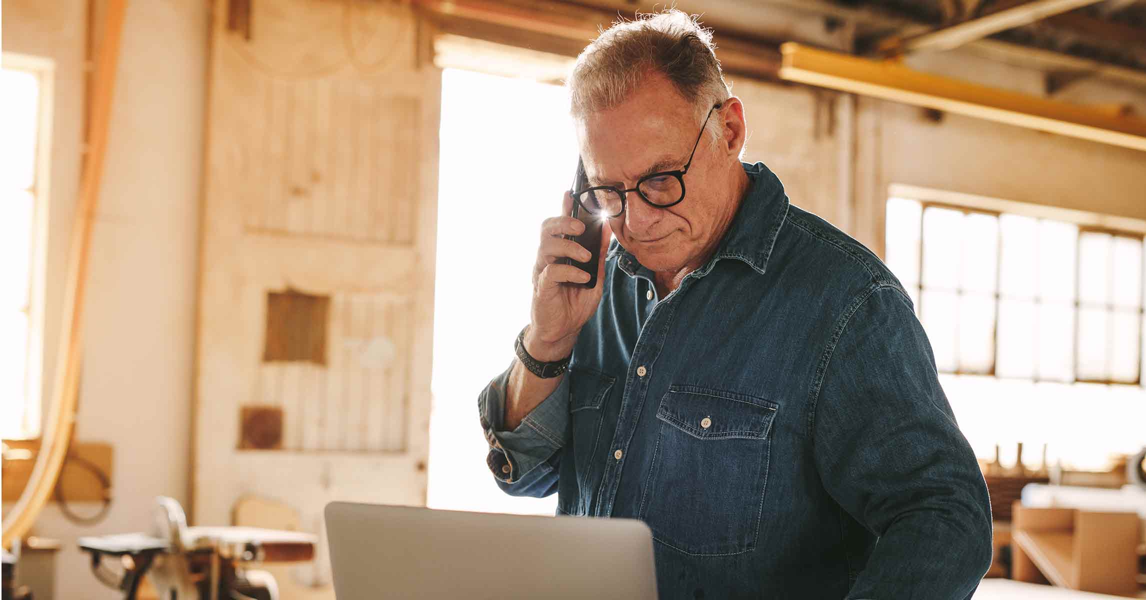 Mature carpenter working on laptop and talking on cell phone