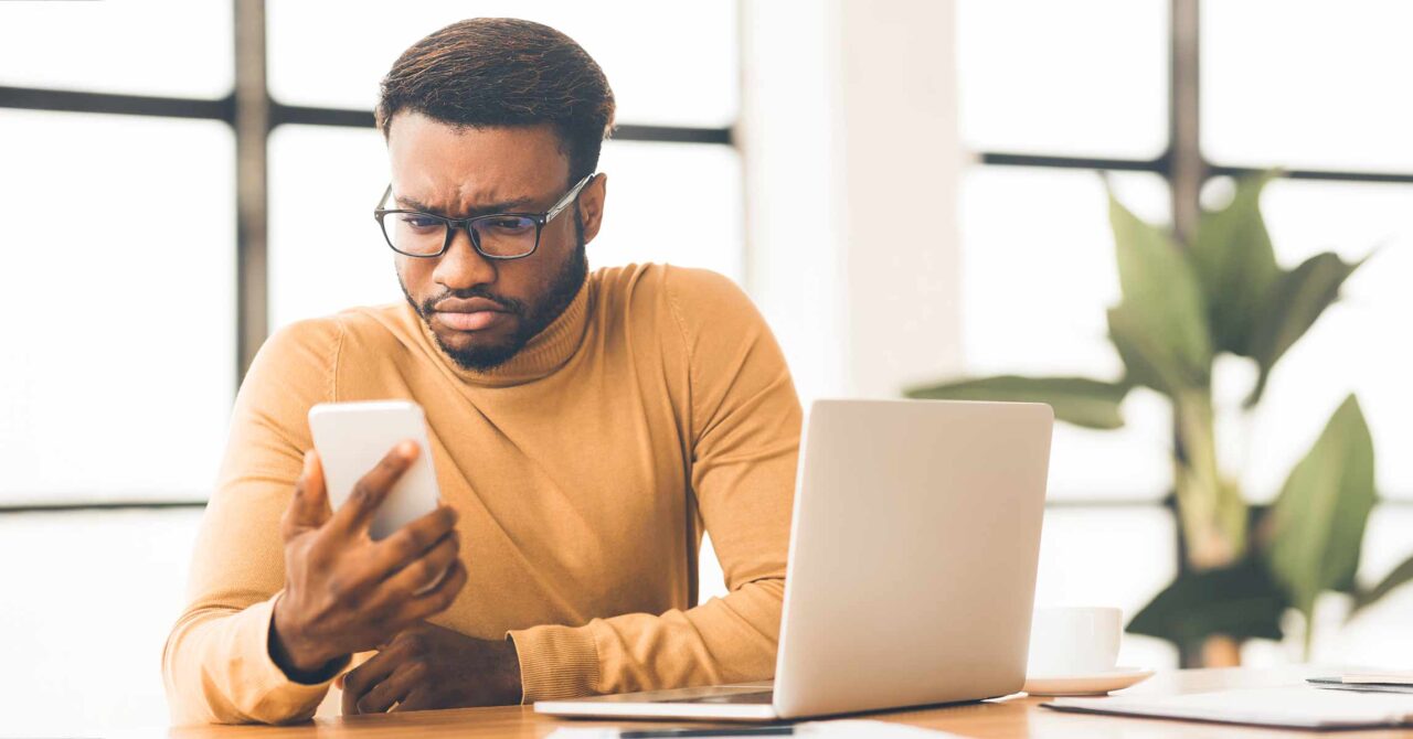 A man at his desk reviewing a message on his phone, pausing to decide if it could be a scam.