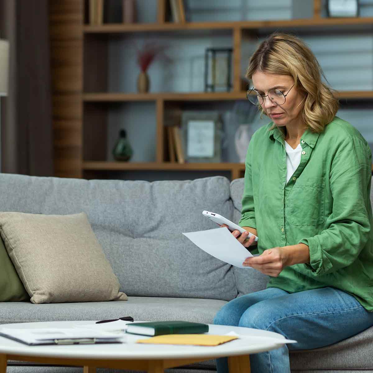 A concerned woman sitting on her couch, reading a bill or notice and calling for help