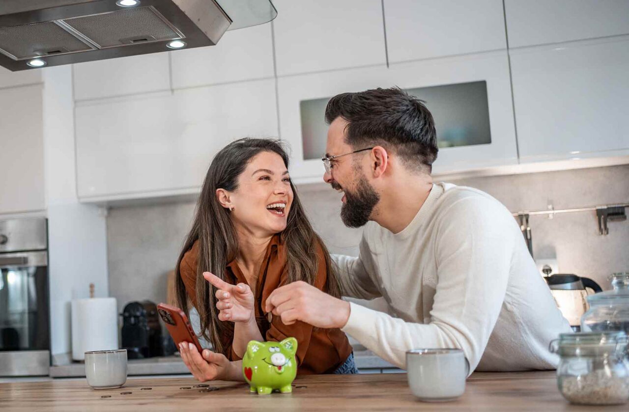 A laughing couple in their kitchen reviewing their finances on a cell phone and dropping a coin into a piggy bank, symbolizing savings and investment
