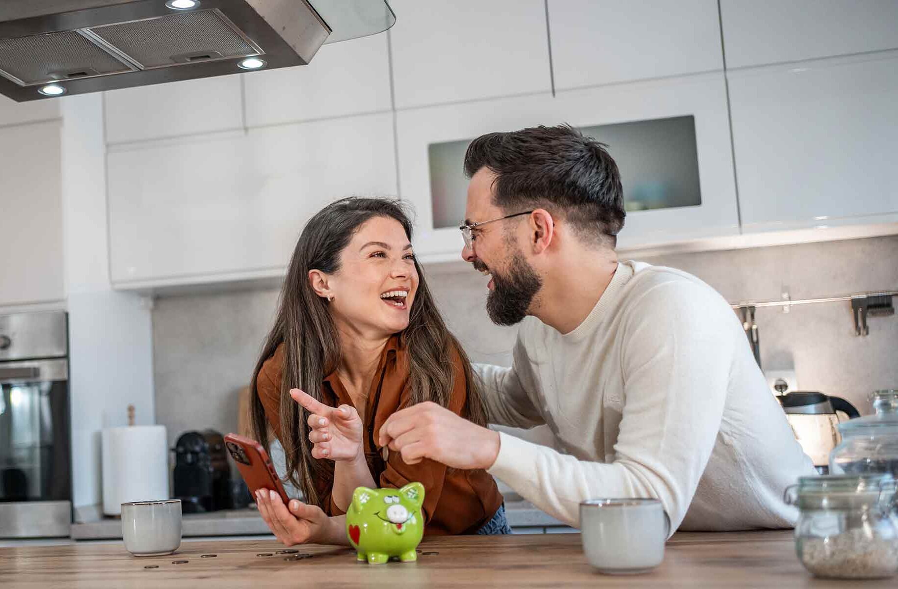 A laughing couple in their kitchen reviewing their finances on a cell phone and dropping a coin into a piggy bank, symbolizing savings and investment