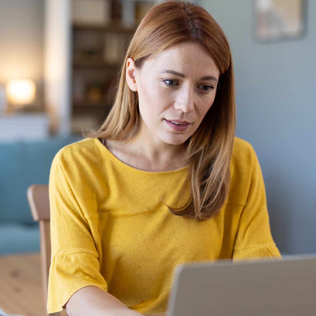 A woman sitting at her kitchen table using a laptop, a skeptical expression on her face