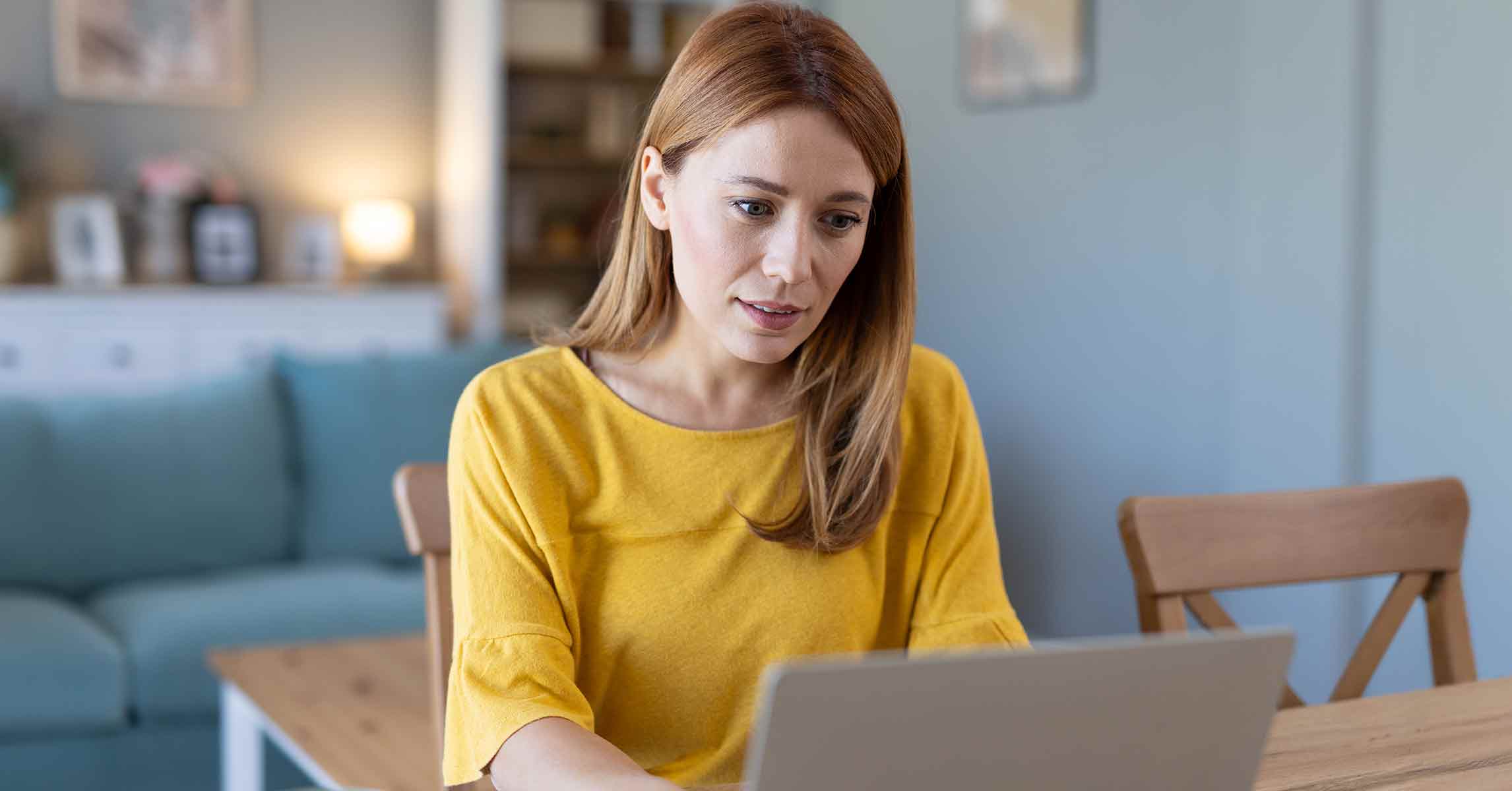 A woman sitting at her kitchen table using a laptop, a skeptical expression on her face