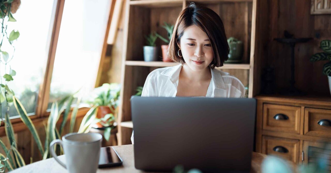 A young woman in her home office, working on her laptop and sipping coffee