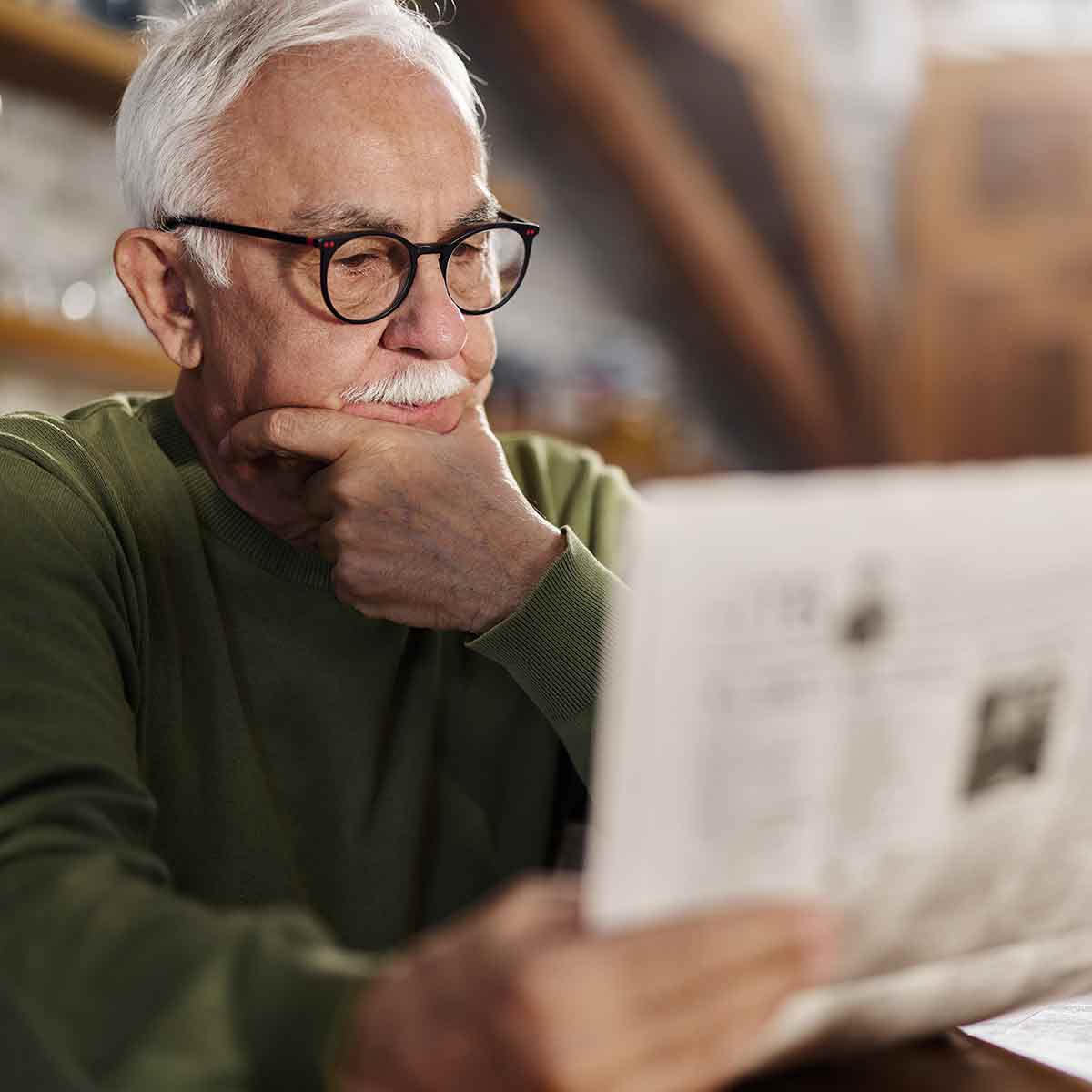 Older man at a kitchen table reading a newspaper and stroking his chin, reflecting uncertainty about financial news