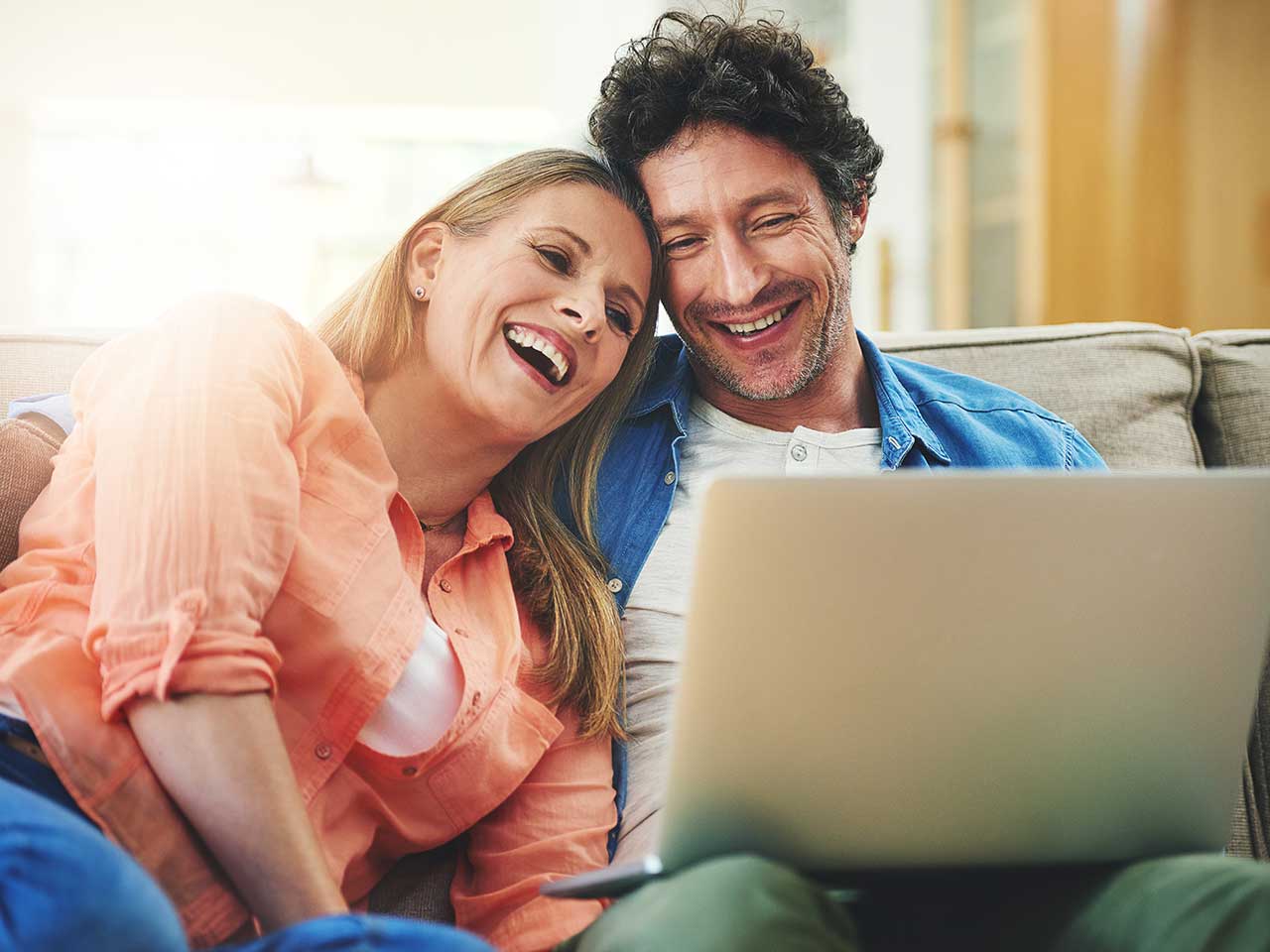 Couple sitting on their couch and smiling while making financial plans on their laptop.